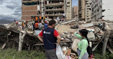 Aid worker and resident stand beside rubble as rescue teams search debris in front of a heavily damaged apartment building.