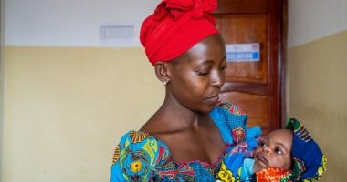 A woman wearing a bright red headscarf and colourful patterned dress cradles her baby in her arms while looking down with care. Wrapped in a vibrant blanket, the baby looks back up at her inside a healthcare facility.