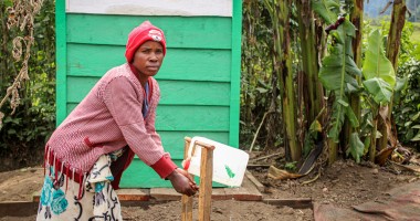 A woman in a pink sweater washing her hands at an outdoor tap in DRC
