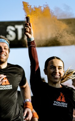 A woman smiles and raises a flare emitting orange powder as she runs under the “START” arch at a Tough Mudder event, surrounded by fellow participants in black Tough Mudder shirts. The atmosphere is energetic and celebratory. 