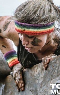 A muddy Tough Mudder participant wearing a rainbow headband and wristbands climbs over a wooden obstacle, showing focus and determination.