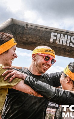 Three muddy Tough Mudder participants celebrate at the finish line, smiling and hugging beneath a large black arch marked “FINISH.” All wear orange headbands and show signs of joy.