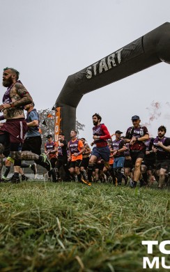A large group of Tough Mudder participants begins their race, running under a black inflatable arch marked “START” on a foggy morning. The ground is grassy, and the scene is energetic with banners and spectators in the background.