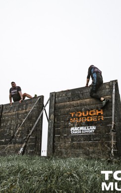 Three Tough Mudder participants climb over tall wooden walls, part of an obstacle course. The walls are branded with Tough Mudder logos. Orange and black flags line the course, and the ground is grassy under a grey sky.
