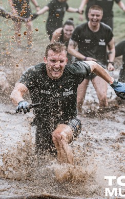 A group of Tough Mudder participants splash through a muddy water obstacle, with one man in front charging ahead with a determined expression. Mud sprays through the air as others laugh and push through behind him.