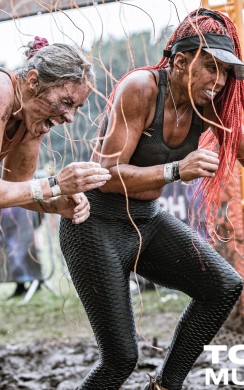 Two Tough Mudder participants grit their teeth as they push through hanging electrical wires in a muddy obstacle. Both are covered in mud and show intense focus, while spectators watch in the background.