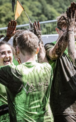 A group of Tough Mudder participants wearing green shirts celebrate with muddy high-fives after completing the course. Their faces are joyful, and they are covered in mud from head to toe.