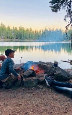 A group of four people, including a child, sit around a small campfire near a calm forest lake at sunset. One person is roasting marshmallows while others relax on logs and rocks. Trees and a red tent are visible in the background, reflecting in the water.