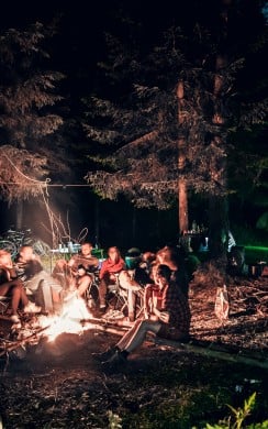 A group of people sit around a glowing campfire at night in a forest clearing, surrounded by tents and tall trees. The scene is warmly lit, with bicycles and camping gear visible in the background.