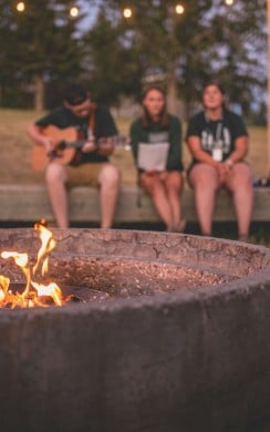 A campfire burns in a large circular fire pit in the foreground, while three people sit on a wooden bench in the background. One plays a guitar, and the other two listen and sing along. String lights are hung above, and the setting is surrounded by trees and open space at dusk.