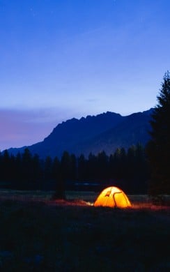 A glowing orange tent is set up in a dark forest clearing at twilight, surrounded by tall trees and mountain silhouettes under a starry sky.