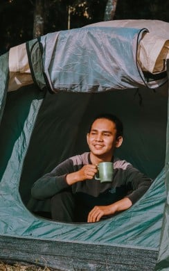 A smiling person sits at the entrance of a green camping tent, holding a green mug. Sunlight shines on their face as they enjoy a warm drink in a forested outdoor setting.