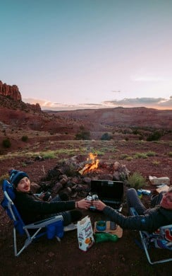 Two people sit in camping chairs in a desert landscape at sunset, toasting drinks by a small campfire. They are surrounded by rocky terrain, with dramatic cliffs in the background and camping gear around them.