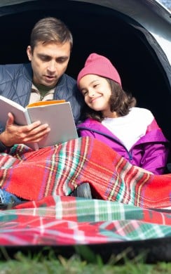A father and daughter sit wrapped in a red tartan blanket inside a tent, reading a book together during a camping trip on a sunny day.