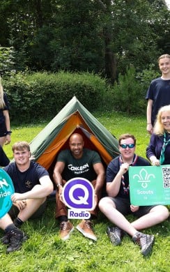 A group of Scouts, Scout leaders, and a Concern Worldwide representative gather around a small tent on a grassy campsite. Several people hold signs featuring the Concern Worldwide logo, Q Radio logo, and Scouts NI logo with a QR code. Surrounded by trees on a sunny day, the group smiles at the camera, highlighting a partnership promoting youth engagement, fundraising, and outdoor adventure.