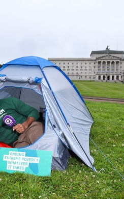A Concern Worldwide supporter sits inside a blue camping tent on the lawn outside Parliament Buildings at Stormont in Belfast. Wearing a Concern Worldwide hoodie and beanie, they hold a Q Radio microphone beside a sign reading “Concern Worldwide – Ending Extreme Poverty, Whatever It Takes.”