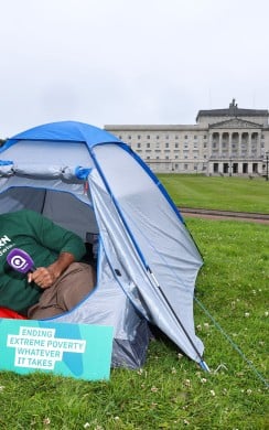 A Concern Worldwide supporter sits inside a blue camping tent on the lawn outside Parliament Buildings at Stormont in Belfast. Wearing a Concern Worldwide hoodie and beanie, they hold a Q Radio microphone beside a sign reading “Concern Worldwide – Ending Extreme Poverty, Whatever It Takes.” 