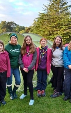 A group of six people, including a Concern Worldwide volunteer and five young Scouts, stand smiling on a grassy campsite in front of several green tents. Surrounded by trees under a bright morning sky, the group wears outdoor clothing and Scout neckerchiefs, highlighting teamwork, youth engagement, and participation in a community camping event.