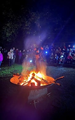 A large group of people gather outdoors at night around a brightly burning campfire. One person stands near the fire with raised arms, appearing to lead the gathering, while others watch from benches and standing areas illuminated by the firelight and handheld torches. Surrounded by trees, the scene conveys a warm sense of community, reflection, and shared experience.