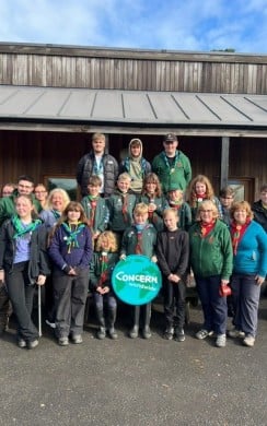 A large group of Scouts, Scout leaders, and volunteers stand together outside a wooden building for a group photo. Several members wear Scout uniforms and neckerchiefs, while others wear outdoor clothing. At the centre of the group, two young people hold a round Concern Worldwide sign. The image highlights a community partnership supporting youth engagement, fundraising, and global solidarity.