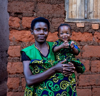 A woman holds a smiling young child outside a home built from mud bricks. Both wear matching green patterned clothing and look toward the camera, while the child smiles and holds a small blue object. A wooden shuttered window is visible behind them.
