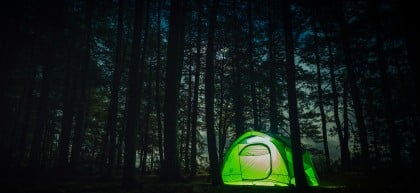 A glowing green tent is set up in a dark forest at night, illuminated from within. Tall trees surround the tent, and faint moonlight filters through the branches, creating a peaceful and secluded atmosphere.