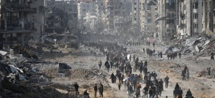 Crowds of people walk through a wide street filled with rubble and debris, flanked by heavily damaged and destroyed buildings in a war-torn city.