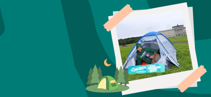 A teal graphic with abstract curved shapes creates a camping-themed backdrop. On the right, a taped, polaroid-style photo shows a person sitting inside a blue tent on a lawn, holding a Q Radio microphone. A Concern Worldwide campaign sign reading "Ending extreme poverty, whatever it takes" is placed in front of the tent, with a large government building visible in the background. Illustrated pine trees, a camping tent and a crescent moon decorate the bottom of the graphic.