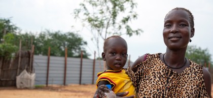 A woman wearing a patterned dress holds a young child dressed in yellow, who is holding a bottle of clean water. They stand outdoors in an open area with trees and fencing in the background, both facing the camera.