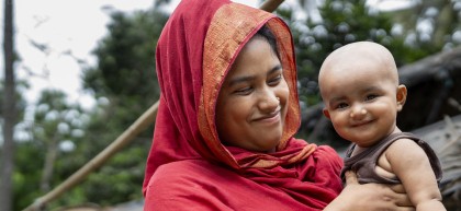 A woman wearing a red headscarf smiles while holding a young child outdoors. The child smiles directly towards the camera, with palm trees and simple homes softly blurred in the background.