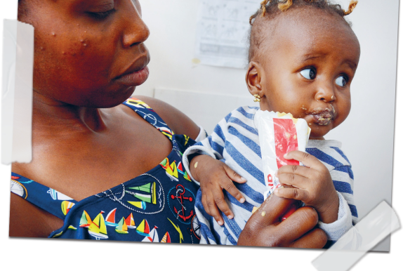 A woman holds a young child in her arms. The child is eating from a sachet of ready-to-use therapeutic food (RUTF) and has food around their mouth. The woman looks down at the child with care. They are indoors, with a white wall and posters in the background.