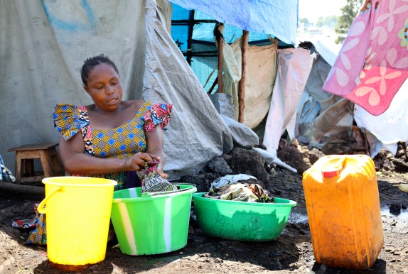 A woman sits in a displacement camp in a colourful dress surrounded by plastic containers.