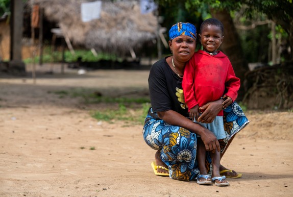 A woman wearing a blue patterned headscarf and patterned wrap skirt crouches beside a smiling young child in a red hoodie on a sandy path. They face the camera together in front of a thatched-roof home, with trees and simple village buildings softly blurred in the background.
