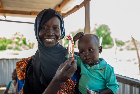 A smiling woman wearing a black headscarf holds a young child while showing a sachet of ready-to-use therapeutic food. The child holds another sachet and looks to one side. They are standing under a covered shelter with a bright outdoor landscape in the background.