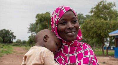 A person wearing a pink and white patterned headscarf holds a child outdoors. In the background, there are trees and a building with a blue roof.