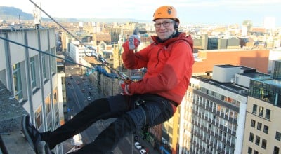 A man wearing a helmet, red jacket, and safety harness abseils down the side of a tall city building, smiling and giving a thumbs-up with one hand. The city skyline stretches into the distance beneath a bright sky.
