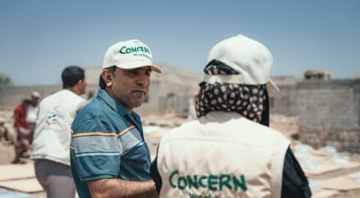Two Concern Worldwide workers talk amid a humanitarian operation. One wears a branded vest and cap, and the other wears a blue shirt with a Concern cap, with tents and unfinished buildings in the background under a clear sky.