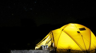 A glowing yellow tent illuminated from within stands in a dark campsite under a star-filled night sky. Hiking boots and trekking poles rest outside the entrance, suggesting a day of adventure.