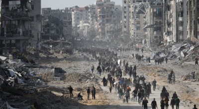 Crowds of people walk through a wide street filled with rubble and debris, flanked by heavily damaged and destroyed buildings in a war-torn city.