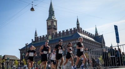 Participants for the Copenhagen Half Marathon run on the course. There are spectators behind barriers either side and a number of runners wearing different coloured running vests including black and white. They run in front of a grand building.