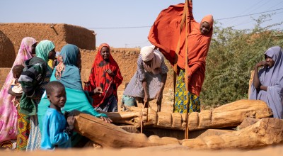 Women collect water from a wooden village well. Her orange hijab billows in the wind as she pulls water up from the well. Behind are orange building and a clear, sunny sky.