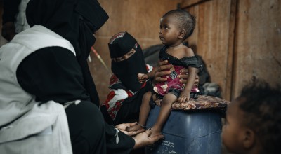 healthcare worker dressed in black clothing with a white vest is examining a small child seated on a blue plastic container. Another person wearing patterned clothing and a black face covering supports the child. The child is wearing a red and black outfit and appears thin, with visible ribs and limbs. The setting looks modest, with a brown wall in the background and another child partially visible in the foreground.