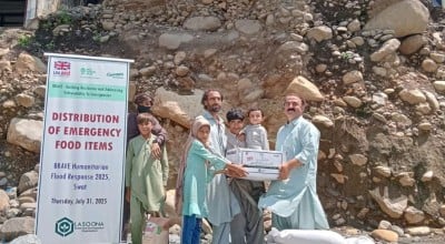 A group of people stand on rocky ground in front of a damaged, partially collapsed house while receiving a box of emergency food supplies. A banner beside them reads “Distribution of Emergency Food Items – BRAVE Humanitarian Flood Response 2025, Swat,” indicating a relief effort supported by aid organisations.