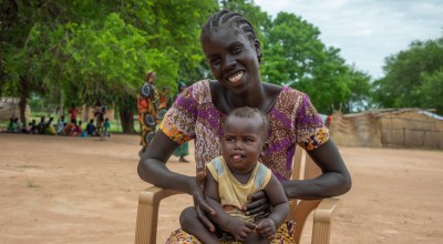A mother sits on a charity with her happy baby on her lap. Behind her are people in colourful clothing and large green trees. 