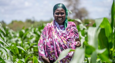 A smiling woman wearing purple stands in a field surrounded by plants.