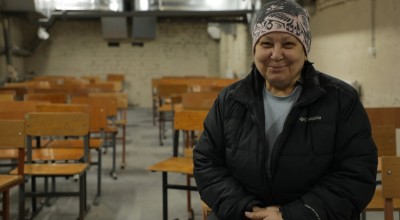 A person wearing a winter jacket and patterned hat sits in a room filled with rows of wooden school chairs. The setting appears to be a basement shelter with exposed brick walls and overhead ventilation ducts.