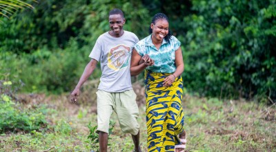  Two people walk, smiling together through a grassy, open area bordered by dense green vegetation. One person wears a grey T‑shirt and light shorts, while the other wears a patterned yellow and blue skirt and a teal top. They hold onto each other’s arms as they move forward across the uneven ground.