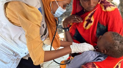 A healthcare worker wearing a face mask and gloves uses a stethoscope to examine a baby held by the child’s mother. The consultation takes place outdoors, with the mother watching attentively as the baby is checked.