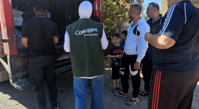 A group of people stand beside the open back of a truck as aid supplies are unloaded. A person wearing a green vest with “Concern Worldwide” on the back helps coordinate the distribution while families wait nearby on a sunny street.