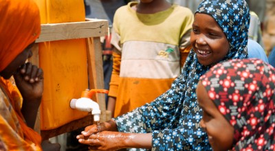 Several children gather around a handwashing station, smiling as one child washes their hands under running water from a yellow container with a tap. The scene shows a shared, outdoor setting with other children watching and waiting their turn.